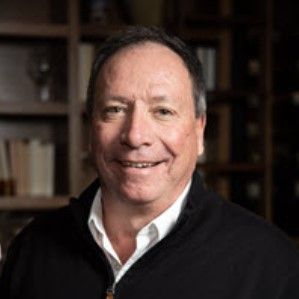 image of a man smiling with greying hair wearing a white collared shirt underneath a black sweater sitting in front of a book shelf