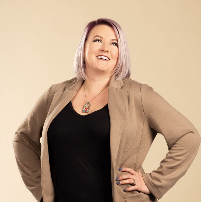 image of a woman with blonde hair smiling and wearing a black top and a tan coat with a large gold necklace