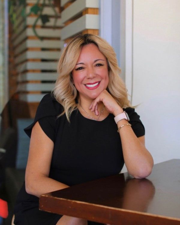 smiling woman sitting at a office desk wearing a black dress and white beaded bracelet