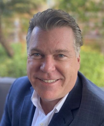 image of smiling man with graying hair wearing a blue suit jacket with a white button up shirt in front of a landscape background