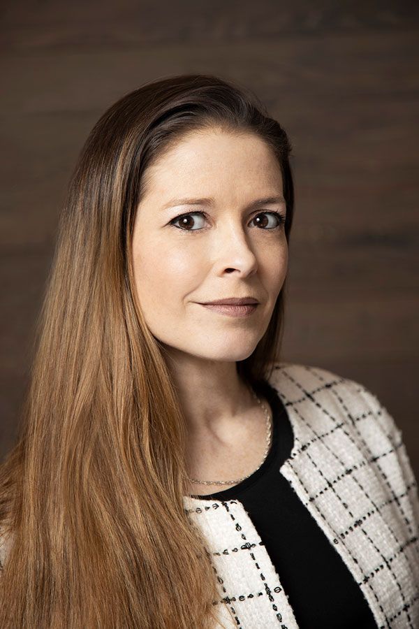 Image of woman with long brown hair posing in front of a brown background wearing a white suit coat with large checks and a black shirt underneath