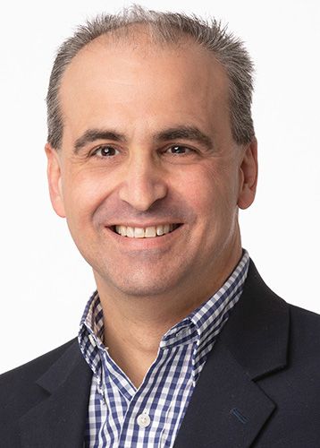 smiling man with salt an pepper hair wearing a blue and white checkered button up shirt and dark blue blazer