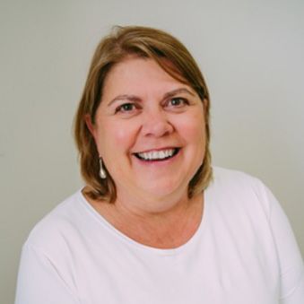 image of smiling woman with light brown hair and wearing a white top in front of a gray background