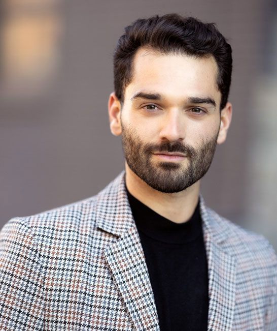 image of a man with dark hair and a dark shadow of a beard wearing a checkered suit coat and a black t-shirt underneath