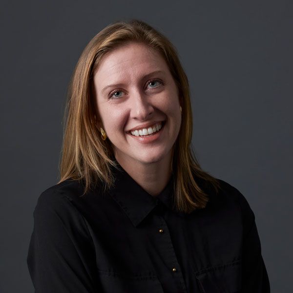 smiling woman with light brown hair posting in front of a dark grey background and wearing a black button up top