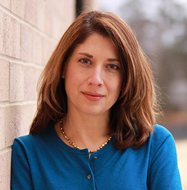 Image of woman with brown hair and a blue top standing against a brick wall