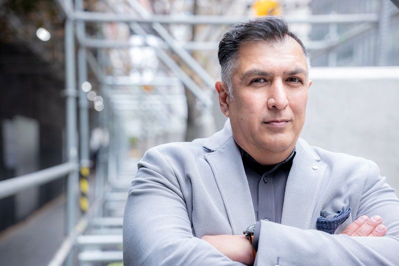 image of man with arms crossed and a slight smile with dark hair speckled with gray and wearing a dark gray t-shirt with a light gray blazer 